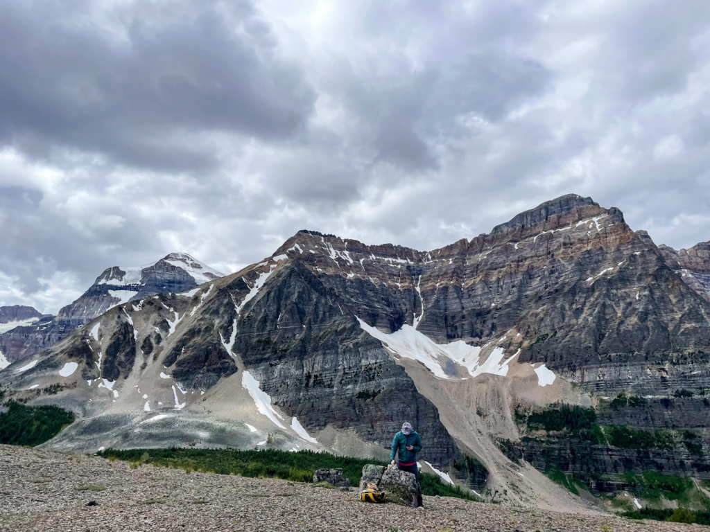 Man looking down near a rock in the foreground, Very large, rocky mountains taking up the most of the background, with a cloudy sky.