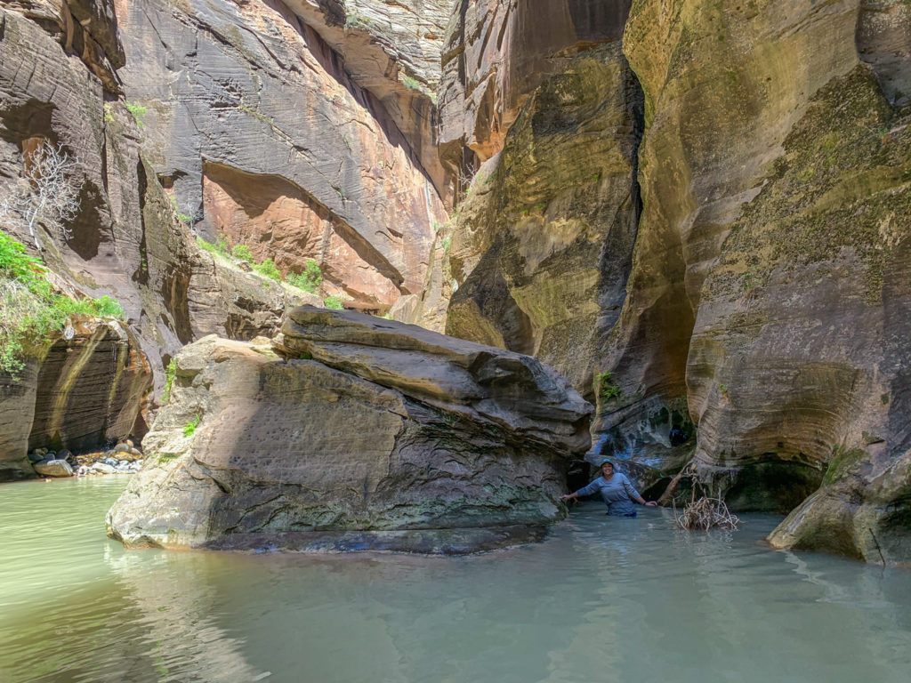 Jack standing next between a large rock in the water and the canyon wall waist deep in water. 