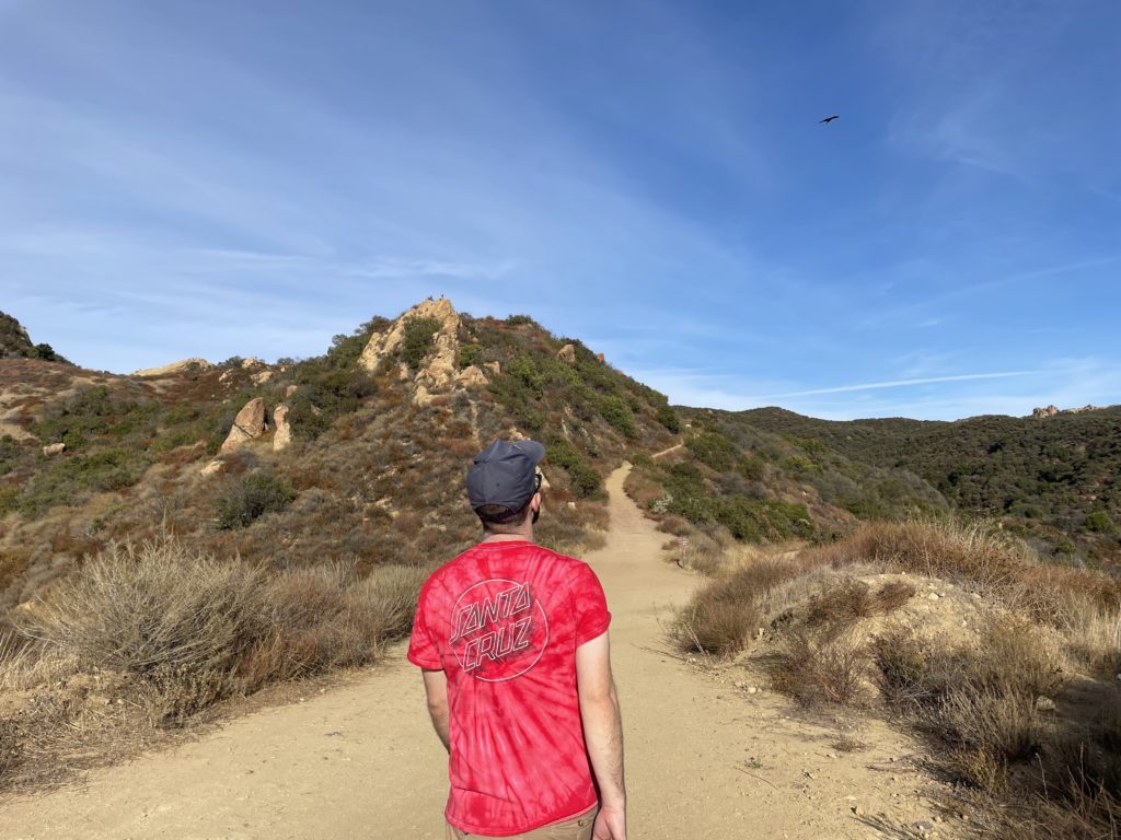 back of man in red santa cruz shirt heading down dusty trail with some dried brush along the hills. 