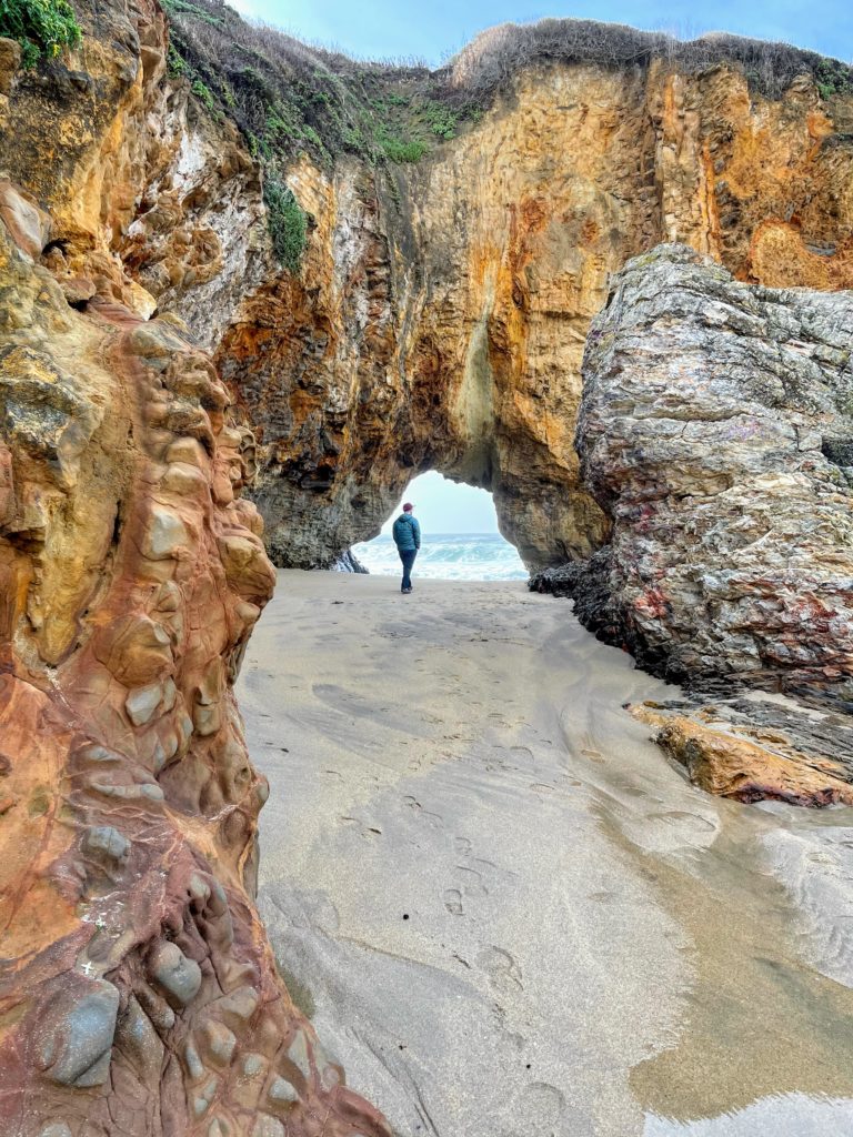 man walking through arch, streaked red and an orange rocks in the foreground, ocean seen through arch. 